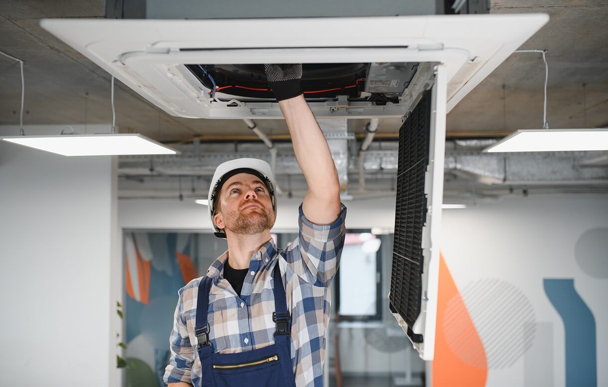 Technician in overalls and a hard hat performing maintenance on a ceiling-mounted HVAC unit in a modern office.
